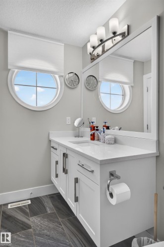 White vanity with a solid-surface countertop and integrated sink, framed rectangular wall mirror, circular window, and charcoal-toned tile flooring - 798 Berg Loop, Leduc, AB 
