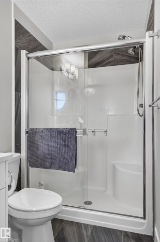 Shower enclosure featuring sliding glass doors, a built-in corner seat, and a handheld showerhead, accented by dark, chevron-patterned tile surrounds and wood-finish floor tiling - 798 Berg Loop, Leduc, AB 