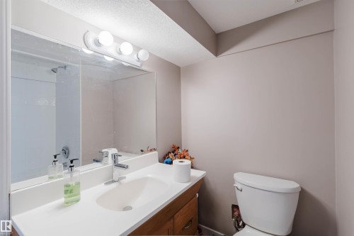 Full-width vanity mirror with four-light wall-mounted fixture, white countertop with integrated sink, wood-finish cabinetry, and white toilet - 225 Heagle Crescent Nw, Edmonton, AB 