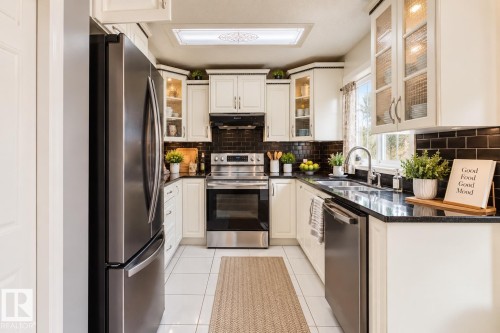 Kitchen featuring cream-colored cabinetry with glass-front uppers, black subway tile backsplash, dark solid-surface countertops, stainless steel appliances, and tile flooring - 225 Heagle Crescent Nw, Edmonton, AB 
