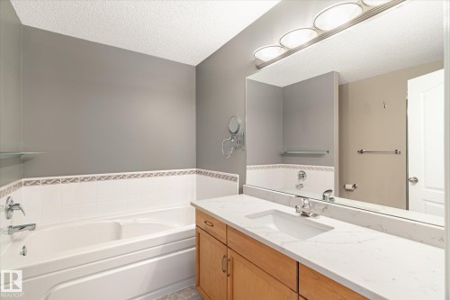 Recessed soaking tub, white wall tiling with decorative trim, wood-finish vanity cabinetry, integrated sink with chrome faucet, and expansive wall-mounted mirror - 2550 Rabbit Hill Road Nw, Edmonton, AB 