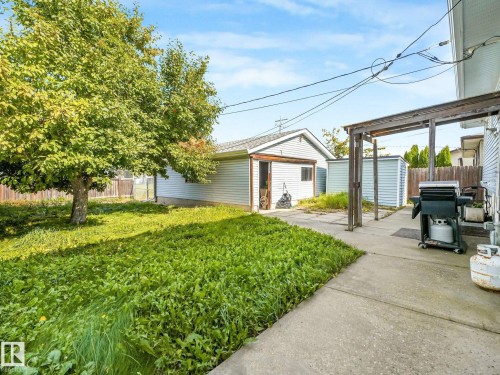 Expansive backyard featuring a mature tree, concrete patio, a detached garage with siding, and two storage sheds - 7520 130 Avenue Nw, Edmonton, AB 