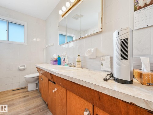 White tiled walls, wood-finish flooring, white vanity countertop, built-in sink, and mounted mirror cabinet with integrated lighting - 7520 130 Avenue Nw, Edmonton, AB 