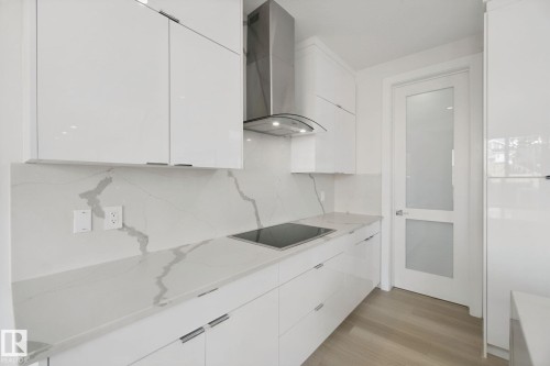 Sleek kitchen featuring white flat-panel cabinetry, a stainless steel range hood, an integrated cooktop, stone countertops with matching backsplash, and wood-finish flooring - 3699 Hummingbird Way Nw, Edmonton, AB 