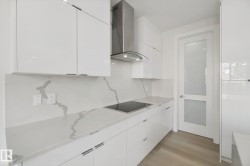 Sleek kitchen featuring white flat-panel cabinetry, a stainless steel range hood, an integrated cooktop, stone countertops with matching backsplash, and wood-finish flooring - 