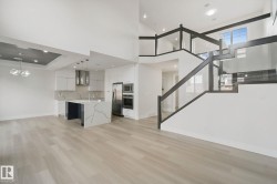 Open-concept design featuring wood-finish flooring, a contemporary kitchen with a waterfall island, stainless steel appliances, and a modern glass and dark-toned staircase - 