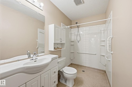 Bathroom featuring a white integrated vanity sink, wall-mounted storage cabinet, full-width vanity mirror, walk-in shower enclosure with integrated shelving, and wall-mounted grab bars - #19 10 Grange Dr, St. Albert, AB 