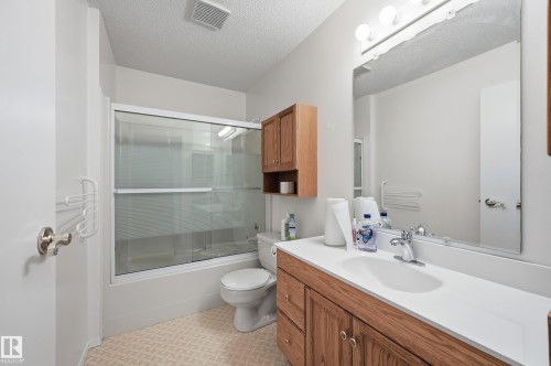 Bathroom featuring wood-finish cabinetry, a white vanity countertop with an integrated sink, a full-width vanity mirror, and a shower-tub combination with sliding glass doors - #19 10 Grange Dr, St. Albert, AB 