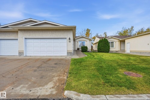 Exterior featuring a light-toned stucco facade, multi-car garage, concrete driveway, and manicured lawn - #19 10 Grange Dr, St. Albert, AB 
