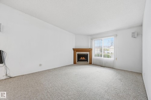 Corner fireplace featuring a wood mantel and tile surround, accompanied by a large window with vertical blinds and wall-to-wall carpeting - #19 10 Grange Dr, St. Albert, AB 