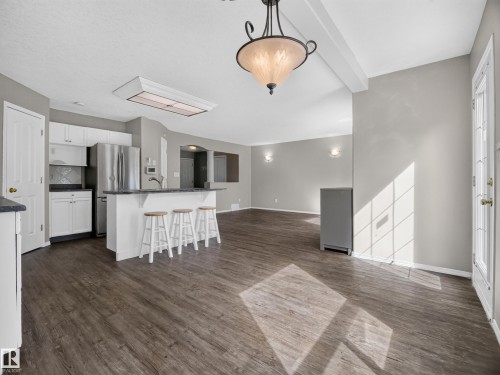 Open-concept living space featuring wood-finish flooring, white cabinetry, a kitchen island with seating, stainless steel appliances, and a decorative overhead light fixture - 14 Wells Court, Leduc, AB 