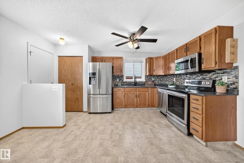 Kitchen featuring wood cabinetry, stainless steel appliances, dark countertops, a mosaic tile backsplash, and light-toned flooring - 14519 32 Street Nw, Edmonton, AB 