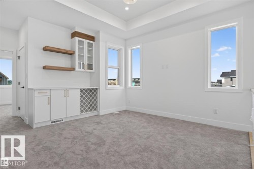 Built-in cabinetry with glass-front uppers and a wine rack, wood-finish floating shelves, light-toned carpeting, and a tray ceiling - 101 Rosa Crescent, St. Albert, AB 