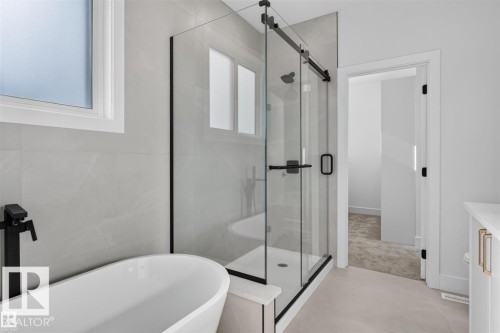 Freestanding soaking tub with a matte black floor-mounted faucet, glass-enclosed shower with black trim, and light-toned wall and floor tiling - 101 Rosa Crescent, St. Albert, AB 