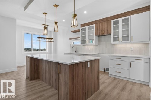 Spacious kitchen featuring a large island with a light-colored countertop, wood-finish flooring, two-tone cabinetry with gold-tone hardware, and pendant lighting - 101 Rosa Crescent, St. Albert, AB 