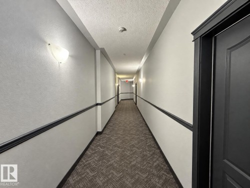 Long hallway with patterned carpet flooring, wall sconce lighting, and contrasting dark trim - #224 3425 19 St Nw, Edmonton, AB 
