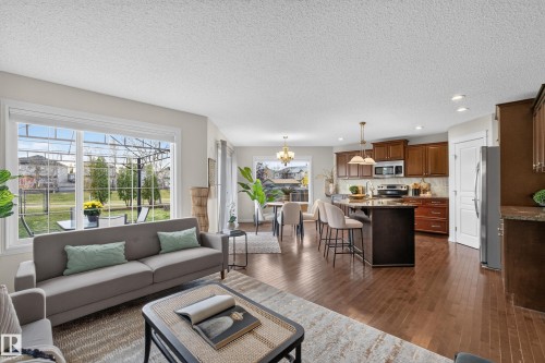 Open-concept interior featuring wood-finish flooring, a central kitchen island with bar seating, custom cabinetry, large picture windows, and recessed lighting - 7 Bremner, Fort Saskatchewan, AB 