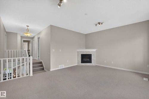 Corner fireplace with stone-finish surround, neutral carpeting, recessed ceiling track lighting, and a white baluster staircase - 7 Bremner, Fort Saskatchewan, AB 