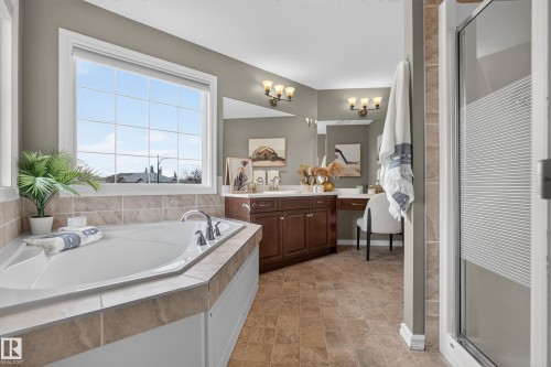 Corner soaking tub with tile surround, wood-finish cabinetry, built-in vanity station, large wall-mounted mirror with multi-bulb lighting, and glass-enclosed shower - 7 Bremner, Fort Saskatchewan, AB 