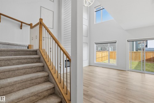 Double-height living area featuring wood-finish flooring, floor-to-ceiling fireplace surround with horizontal detailing, staircase with wooden handrail, and sliding glass doors - 2045 209A St Nw, Edmonton, AB 
