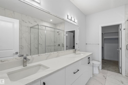 Dual-sink vanity with white cabinetry, light-toned stone-finish countertops, large vanity mirror, walk-in shower with glass enclosure, and large-format tile flooring - 2045 209A St Nw, Edmonton, AB 