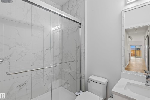 Full-height wall tiling with a vein pattern, glass-enclosed shower with metal hardware, vanity with a rectangular sink, stone-finish countertop, and decorative mirror frame - 2045 209A St Nw, Edmonton, AB 