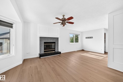 Spacious room featuring wood-finish flooring, a contemporary fireplace with dark tile surround and hearth, multiple windows, and a ceiling fan - 3924 26A Avenue Nw, Edmonton, AB 