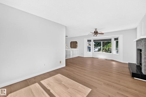Wood-finish flooring throughout, featuring a stone fireplace with a black hearth, a bay window, and a ceiling fan with integrated lighting - 3924 26A Avenue Nw, Edmonton, AB 