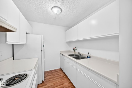 Compact kitchen featuring wood-finish flooring, white cabinetry, light-toned countertops, a double basin sink, and a refrigerator - #212 11218 80 St Nw, Edmonton, AB 