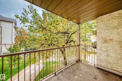Private balcony with a metal railing, textured exterior wall, and overhead paneling, offering views of mature trees and a paved walkway - #212 11218 80 St Nw, Edmonton, AB 