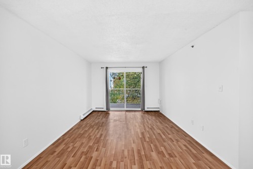 Wood-finish flooring throughout, bright white walls, and a sliding glass door leading to a balcony with a black metal railing - #212 11218 80 St Nw, Edmonton, AB 