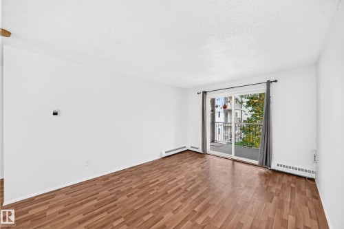Wood-finish flooring, white painted walls, and a sliding glass door to a private balcony with a metal railing - #212 11218 80 St Nw, Edmonton, AB 