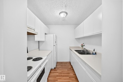 Galley kitchen featuring white cabinetry, light countertops, wood-finish flooring, and a double-basin sink - #212 11218 80 St Nw, Edmonton, AB 