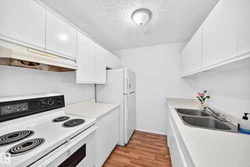 Galley kitchen featuring wood-finish flooring, white cabinetry, light-colored countertops, a double basin sink, and a built-in electric range - #212 11218 80 St Nw, Edmonton, AB 