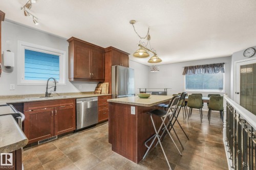 Kitchen featuring stained wood cabinetry, stone-finish countertops, stainless steel appliances, central island with integrated seating space, and tile flooring - 9617 151 Street Nw, Edmonton, AB 