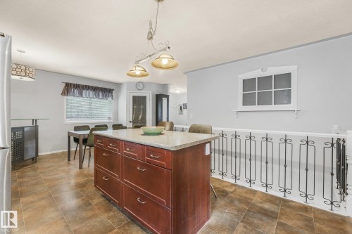 Kitchen featuring a central island with deep-toned cabinetry and a stone-finish countertop, decorative wrought-iron railing, ceramic tile flooring, and multi-light hanging pendant fixtures - 9617 151 Street Nw, Edmonton, AB 