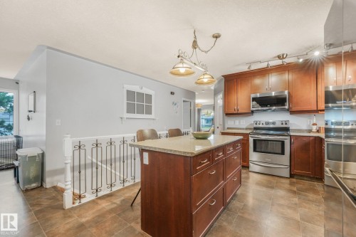 Kitchen featuring wood cabinetry, central island with stone-surface countertop, stainless steel appliances, tile flooring, and decorative metal stair railing - 9617 151 Street Nw, Edmonton, AB 