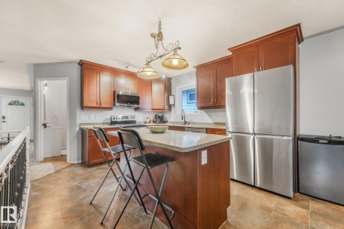 Kitchen featuring wood cabinetry, stone-finish countertops, stainless steel appliances, a center island, and tile flooring - 9617 151 Street Nw, Edmonton, AB 