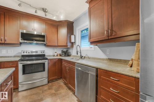 Kitchen featuring stained wood cabinetry, stainless steel appliances, solid-surface countertops, dual-basin sink with a matte black faucet, and track lighting - 9617 151 Street Nw, Edmonton, AB 
