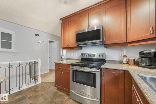 Kitchen featuring stained wood cabinetry, stainless steel appliances, stone-finish countertops, tile flooring, and decorative metal balusters - 9617 151 Street Nw, Edmonton, AB 