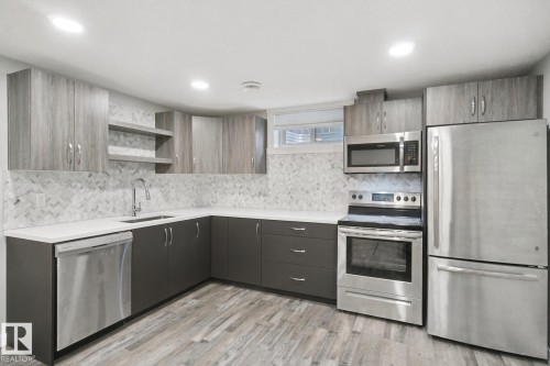 Modern kitchen featuring stainless steel appliances, a herringbone tile backsplash, gray base cabinetry, wood-finish upper cabinets, and light-toned countertops - 13212 73 Street Nw, Edmonton, AB 