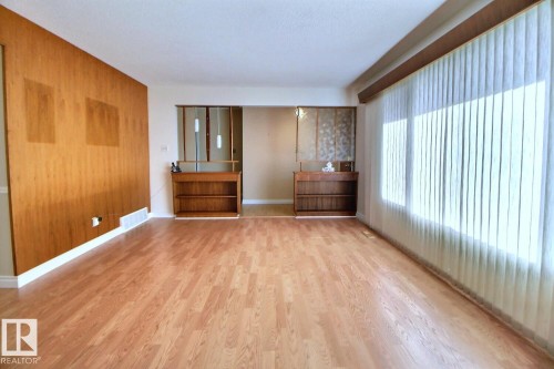 Wood-finish flooring throughout, featuring a wood-paneled accent wall, a wide window with vertical blinds, and built-in shelving units - 4420 117A Street Nw, Edmonton, AB 