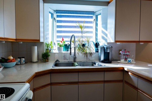 Bay window with horizontal blinds, stainless steel sink with gooseneck faucet, light-toned countertops, and white cabinetry with wood trim - 11119 137 Avenue Nw, Edmonton, AB 