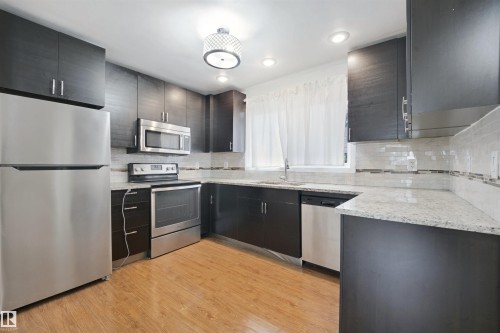 Kitchen featuring dark-toned cabinetry, stainless steel appliances, light-toned countertops, tile backsplash, and wood-finish flooring - 13420 117 Avenue Nw, Edmonton, AB 