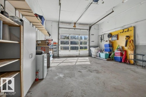 Concrete flooring in a spacious garage featuring a paneled overhead door with integrated windows, built-in wooden shelving, and a wall-mounted storage system - #74 22555 Twp Road 530, Rural Strathcona County, AB 