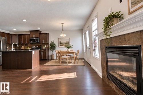 Open-concept living space with wood-finish flooring, a kitchen island, dark wood cabinetry, a built-in fireplace with tile surround, and multiple windows - 5205 168 Avenue Nw, Edmonton, AB 