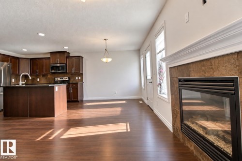 Open-concept living space featuring wood-finish flooring, a built-in electric fireplace with tile surround, a kitchen island with sink, dark wood cabinetry, and stainless steel appliances - 5205 168 Avenue Nw, Edmonton, AB 