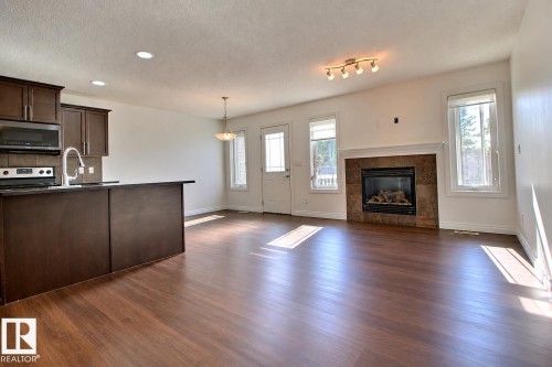 Wood-finish flooring throughout, dark wood-finish cabinetry, stainless steel appliances, a built-in fireplace with a tile surround and mantel, and an overhead track lighting fixture - 5205 168 Avenue Nw, Edmonton, AB 