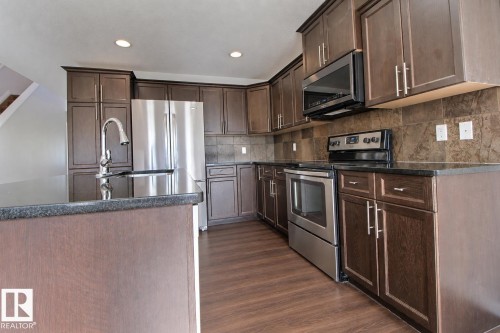 Kitchen featuring dark wood-finish cabinetry, stainless steel appliances, dark countertops, a large kitchen island with an integrated sink, and wood-finish flooring - 5205 168 Avenue Nw, Edmonton, AB 