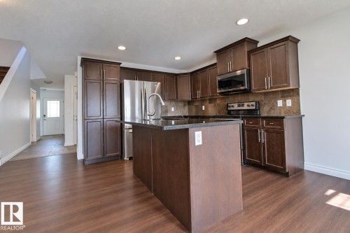 Kitchen featuring wood-finish flooring, dark wood cabinetry, a central island with a dark countertop, stainless steel appliances, and recessed lighting - 5205 168 Avenue Nw, Edmonton, AB 